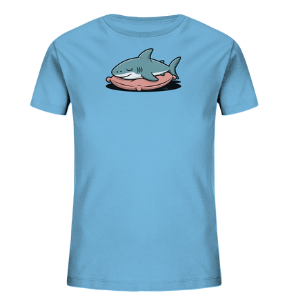 Onlineshop Shirts fuer Kinder - Das Bild zeigt ein hellblaues T-Shirt fuer Kinder mit einem niedlichen Motiv auf der Vorderseite. Darauf ist ein graublauer Cartoon-Hai zu sehen, der entspannt auf einem rosafarbenen Kissen liegt und die Augen geschlossen hat. Das Design wirkt verspielt und friedlich, ideal fuer Kinder.