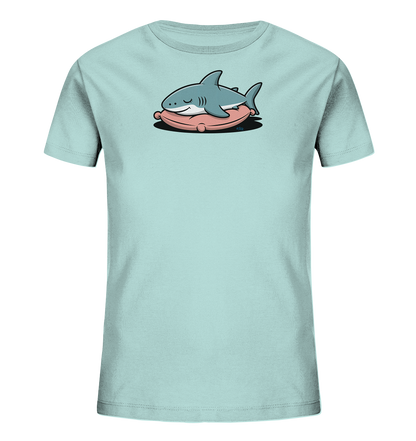 Onlineshop Shirts fuer Kinder - Das Bild zeigt ein hellblaues Kinder-T-Shirt mit einem niedlichen Cartoon-Hai auf der Vorderseite. Der Hai ist in einem ruhigen Zustand dargestellt, mit geschlossenen Augen und einem leichten Laecheln. Er liegt bequem auf einem rosafarbenen Kissen, was dem Motiv eine entspannte und verspielte Note verleiht. Das Design ist einfach und ansprechend fuer Kinder.