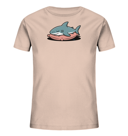 Onlineshop Shirts fuer Kinder - Das Bild zeigt ein hellbeiges Kinder-T-Shirt mit einem niedlichen Aufdruck. Ein cartoonhafter, hellblauer Hai liegt schlafend auf einem rosafarbenen Kissen. Die Augen des Hais sind geschlossen und sein Ausdruck ist friedlich. Das Design ist verspielt und wirkt beruhigend, ideal fuer ein Kinderoutfit.