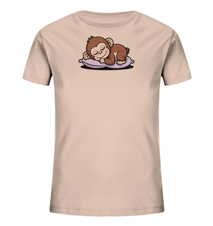 Onlineshop Shirts fuer Kinder - Das Bild zeigt ein hellbeiges Kinder-T-Shirt mit einem niedlichen Tiermotiv auf der Vorderseite. Ein kleiner, brauner Affe liegt schlafend und laechelnd auf einem violetten Kissen. Das Motiv ist detailreich gestaltet und verleiht dem Shirt einen verspielten und friedlichen Charakter. Das T-Shirt ist schlicht gehalten und laesst das Design im Vordergrund stehen.