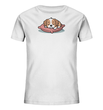 Onlineshop Shirts fuer Kinder - Das Bild zeigt ein weisses Kinder-T-Shirt mit einem niedlichen Tiermotiv auf der Vorderseite. Ein kleiner, braun-weisser Welpe schlaeft friedlich mit geschlossenen Augen auf einem rosafarbenen Kissen. Die Darstellung ist im Cartoon-Stil gehalten und wirkt sehr beruhigend. Das Design verleiht dem Shirt einen liebevollen und gemuetlichen Charakter.