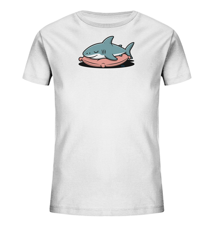 Onlineshop Shirts fuer Kinder - Das Bild zeigt ein weisses Kinder-T-Shirt mit einem niedlichen Aufdruck. Ein hellblauer Cartoon-Hai liegt gemuetlich auf einem rosafarbenen, kissenartigen Objekt. Der Hai hat geschlossene Augen und einen zufriedenen Ausdruck, was eine ruhige und verspielte Szene darstellt. Das Design ist kindgerecht gestaltet.