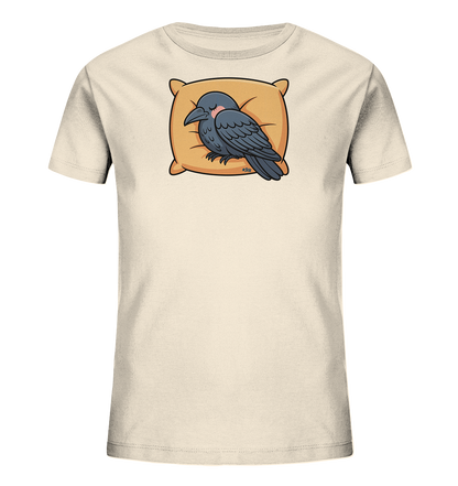 Onlineshop Shirts fuer Kinder - Das Bild zeigt ein hellbeiges T-Shirt fuer Kinder. Die Vorderseite ziert eine charmante Cartoon-Illustration eines dunkelgrauen Raben. Der Rabe liegt friedlich schlafend auf einem hellorangen Kissen und hat leicht geroetete Wangen. Dieses Design verleiht dem Shirt einen ruhigen und liebenswerten Charakter.