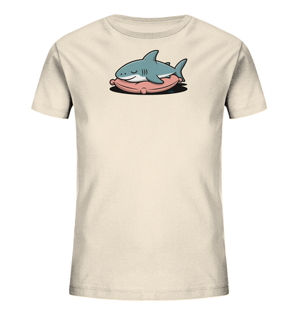 Onlineshop Shirts fuer Kinder - Das Bild zeigt ein hellbeiges Kinder-T-Shirt. Auf der Vorderseite ist ein niedlicher, blauer Cartoon-Hai abgebildet, der entspannt auf einem rosa Kissen liegt. Der Hai hat geschlossene Augen und einen kleinen, freundlichen Gesichtsausdruck. Das Design wirkt ruhig und verspielt.