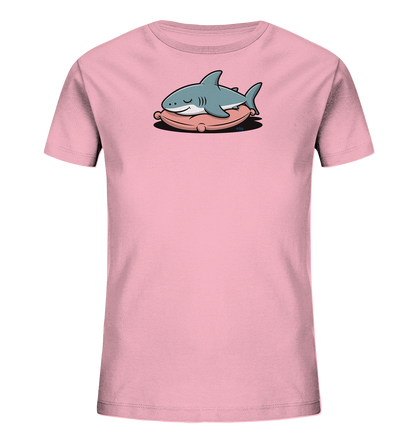 Onlineshop Shirts fuer Kinder - Das Bild zeigt ein hellrosa T-Shirt fuer Kinder. Auf der Vorderseite ist ein niedlicher, hellblauer Cartoon-Hai abgebildet, der friedlich auf einem rosafarbenen Kissen liegt. Der Hai hat geschlossene Augen und ein sanftes Laecheln, was dem Motiv einen beruhigenden Charakter verleiht. Das Design ist verspielt und ideal fuer Kinder.
