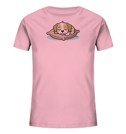 Onlineshop Shirts fuer Kinder - Das Bild zeigt ein hellrosa T-Shirt fuer Kinder. Auf der Vorderseite ist eine niedliche Illustration eines schlafenden Welpen zu sehen, der auf einem rosa Kissen liegt. Der Welpe hat braunes und beiges Fell und seine Augen sind geschlossen, was einen ruhigen und friedlichen Eindruck vermittelt. Das Design ist verspielt und ideal fuer ein Kinderoutfit.